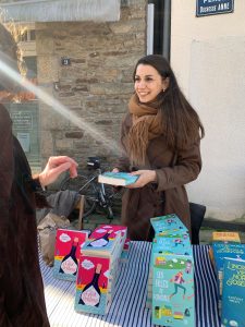 On voit l'autrice Angéline Michel qui tient son dernier roman Les filles de Kinsale dans les mains. Elle porte un manteau en laine marron et a les cheveux détachés. Elle sourit. Elle est en séance de dédicace, derrière une table remplie de ses ouvrages.
